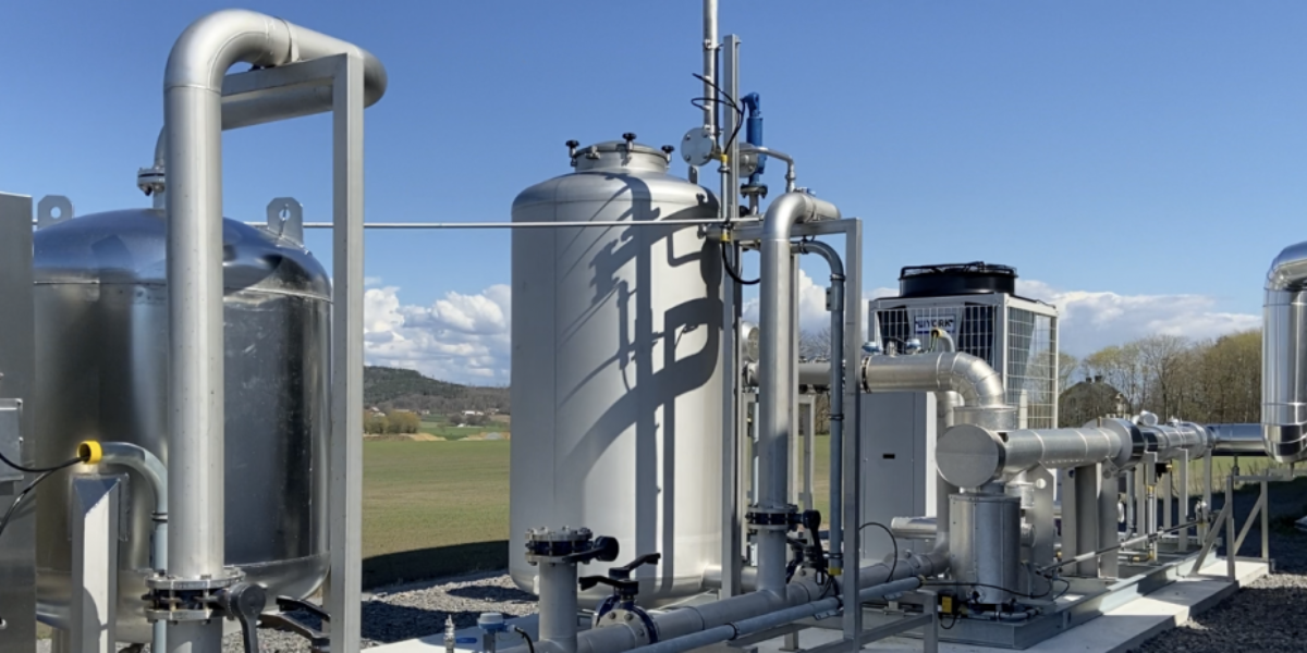 Industrianläggning för biogas med stora silverfärgade tankar och rörledningar som löper längs marken. Utrustningen står utomhus på grus intill en byggnad, med öppna fält och blå himmel i bakgrunden.