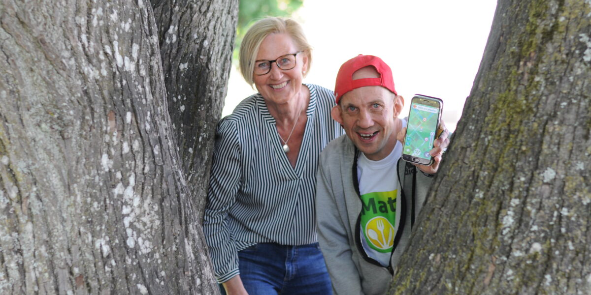 En leende kvinna och en man i röd keps står mellan två stora trädstammar utomhus; mannen håller upp en mobil med en kartapp och bär en tröja med texten “Mat”.