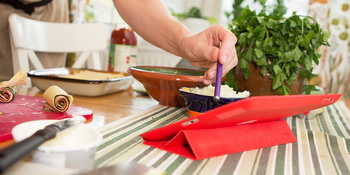 En person rör om i en liten skål med finhackad lök med en lila sked, vid ett köksbord med surfplatta i rött fodral, rullade skivor på skärbräda, grytor och en kruka färska örter.