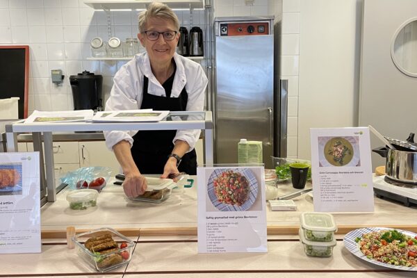 Kock i vitt och svart förkläde förbereder mat vid en köksbänk och ler mot kameran. Framför står upplagda rätter, matlådor och receptblad med rubrikerna ”Åkerburgare med ärtfärs”, ”Saftig grynsallad med gröna åkerbönor” och ”Grön currysoppa åkerböna och broccoli”.