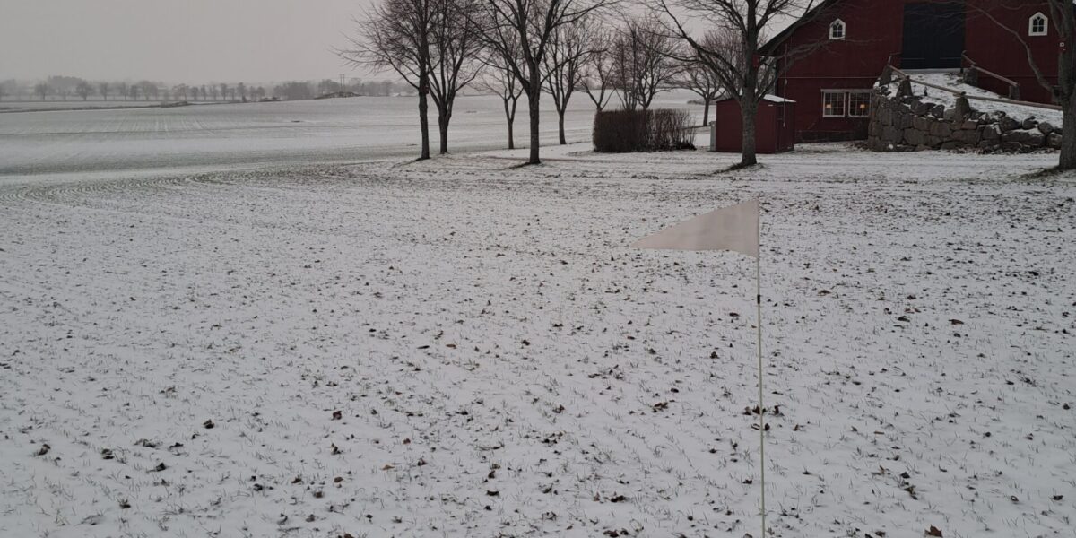 Vit triangelflagga på en smal pinne står i en snötäckt åker, som en markering i förgrunden. I bakgrunden syns nakna träd och en röd ladugård vid en stenmur under grå vinterhimmel.