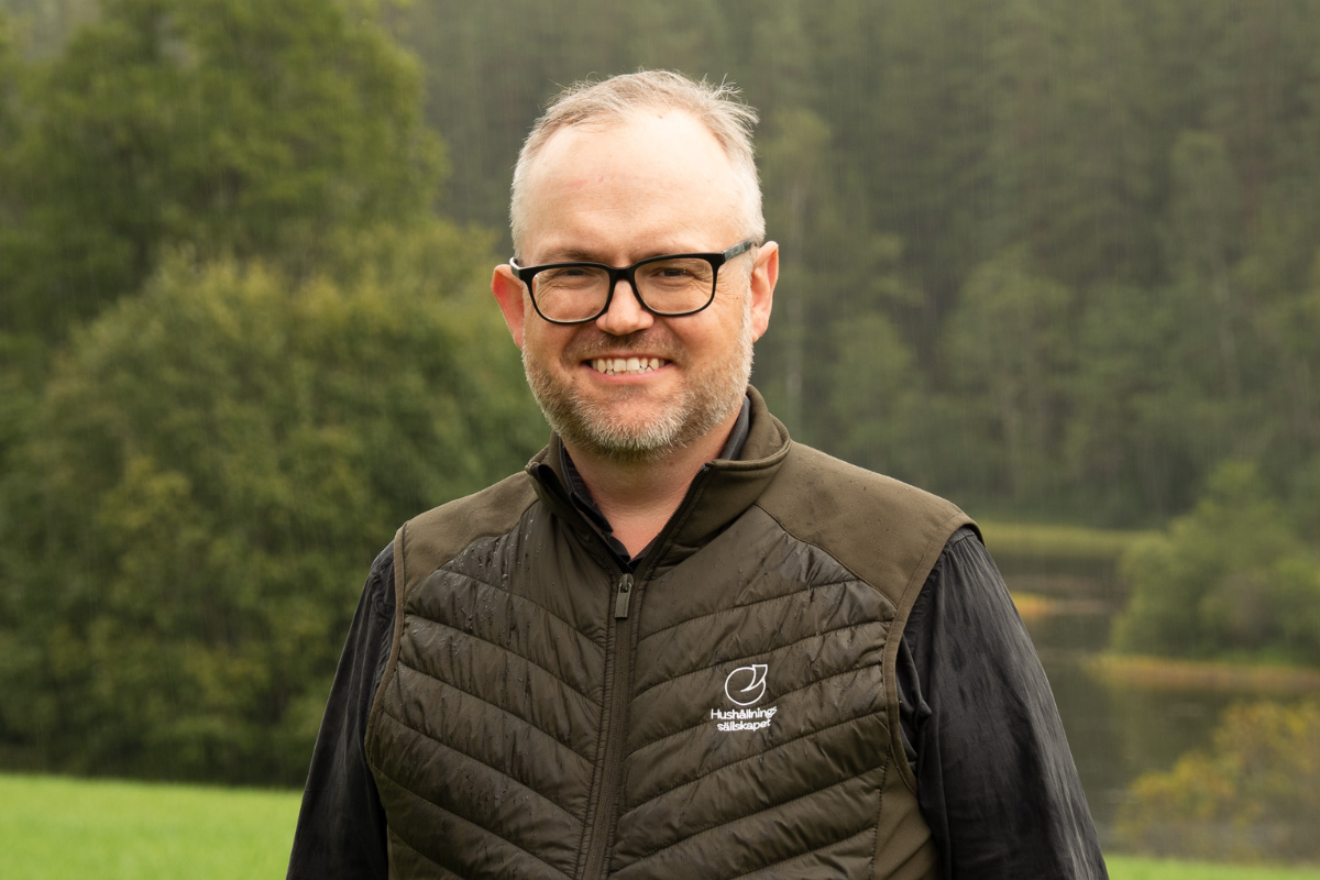 Leende man med svart glasögon står utomhus i lätt regn, iklädd mörk skjorta och quiltad väst med texten ”Hushållningssällskapet”. I bakgrunden syns grönskande skog och en sjö i dimmigt, lugnt landskap.