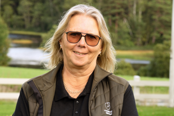 Kvinna med axellångt blont hår och solglasögon står och ler mot kameran utomhus. Hon bär mörk pikétröja och grön väst med logotypen ”HundHälsa Sverige”, med sjö och skog i bakgrunden.