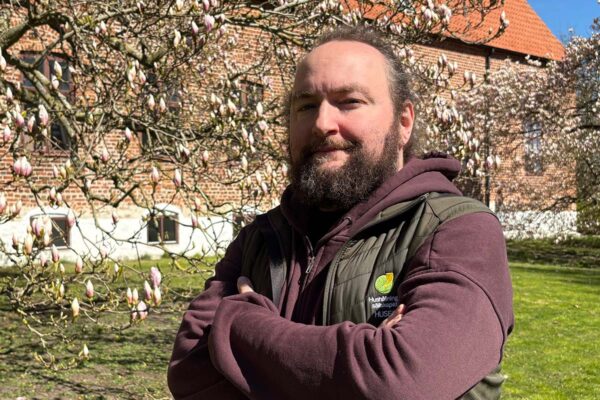 Skäggig man står med armarna i kors och tittar mot kameran, utomhus framför ett blommande magnoliaträd. I bakgrunden syns en tegelbyggnad med röda tak. På västen står ”Hushållningssällskapet” och ”HUSE”.