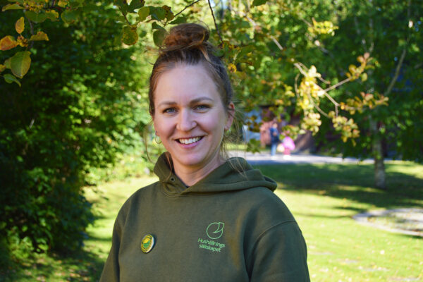 Leende kvinna i grön huvtröja står utomhus under trädgrenar och tittar mot kameran. På tröjan syns texten ”Hushållningssällskapet” och en liten rund pin. Solig parkmiljö i bakgrunden.