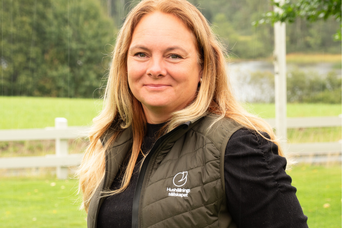 Leende kvinna med långt blont hår tittar mot kameran och står utomhus i lätt regn. Hon bär mörk tröja och grön väst med texten ”Hushållningssällskapet”, med grön gräsmatta, vitt staket och träd i bakgrunden.