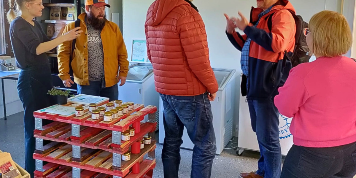 Fyra personer samtalar i en liten butikslokal; en äldre man i röd jacka gestikulerar medan de andra lyssnar. I förgrunden står en röd lastpall med små glasburkar, och bakom syns frysboxar och hyllor.