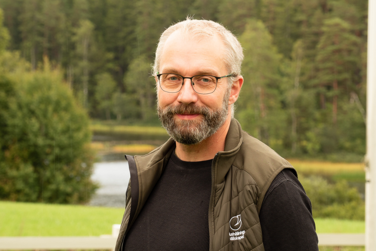 Man med grått hår, skägg och glasögon står utomhus och ler mot kameran, klädd i svart tröja och grön väst med texten ”Hushållnings sällskapet”. I bakgrunden syns en sjö och tät skog.