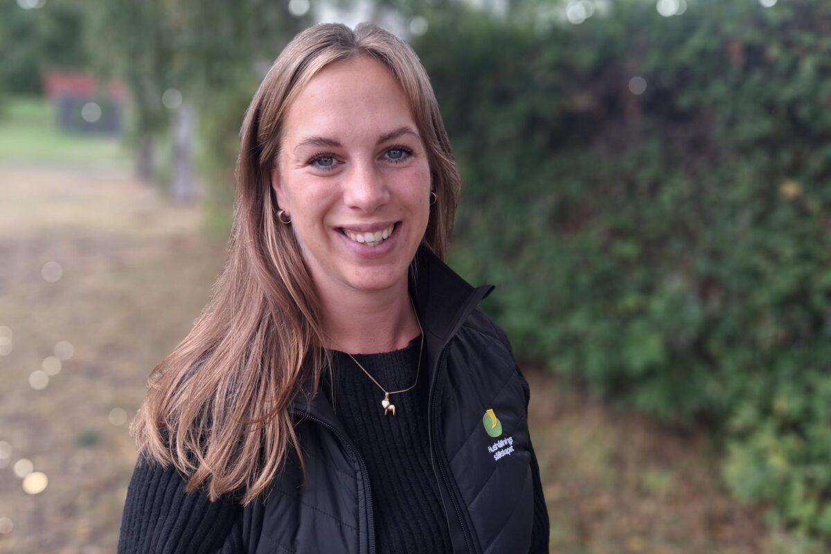 Leende kvinna med långt brunt hår står utomhus framför suddiga träd och en grusväg, tittar mot kameran. Hon bär svart tröja och svart väst med texten ”Hushållningssällskapet”.