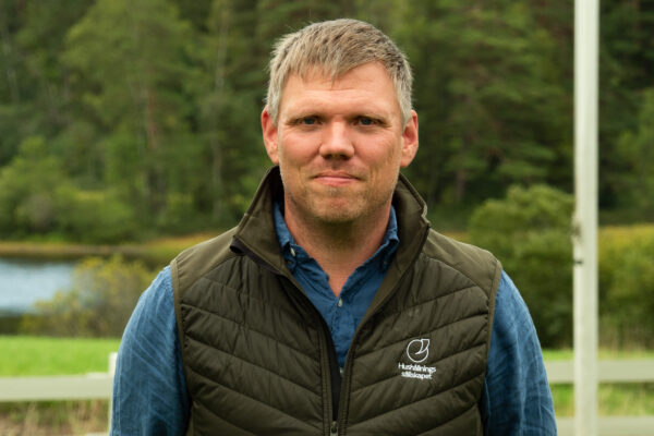 Man med kort, ljusbrunt hår står och tittar in i kameran utomhus, iklädd blå skjorta och grön vadderad väst med texten “Hushållnings sällskapet”, med skog och sjö suddigt i bakgrunden.
