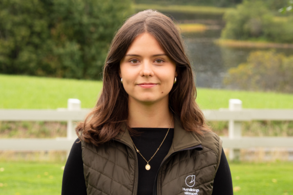 Ung kvinna med brunt, axellångt hår står utomhus och ser in i kameran med ett svagt leende. Hon bär svart tröja, olivgrön väst med texten ”Hushållningssällskapet”, samt guldsmycken; grön park och vit staket bakom.