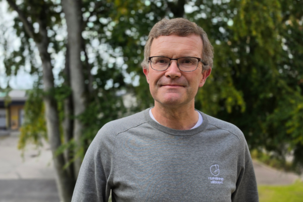 Man med glasögon står och tittar in i kameran med ett svagt leende. Han bär grå tröja med loggan och texten ”Hushållningssällskapet”. I bakgrunden syns grönskande träd och en suddig gårdsmiljö.