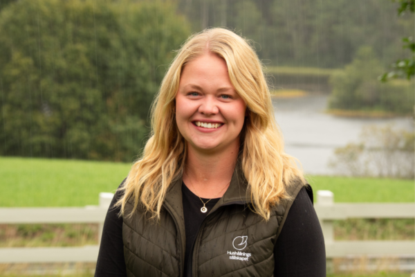Leende kvinna med blont hår står utomhus i lätt regn och tittar mot kameran. Hon bär svart tröja och mörkgrön väst med loggan och texten ”Hushållningssällskapet”, med sjö och skog i bakgrunden.