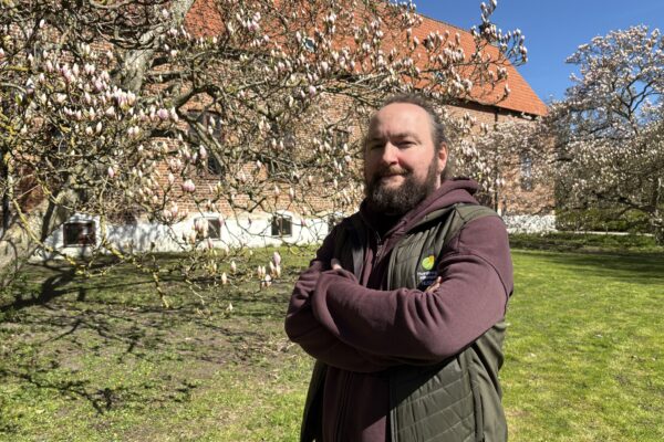 Skäggig man står med armarna i kors och tittar mot kameran, framför ett magnoliaträd fullt av rosa knoppar. I bakgrunden syns en tegelbyggnad med rött tegeltak och blå himmel.
