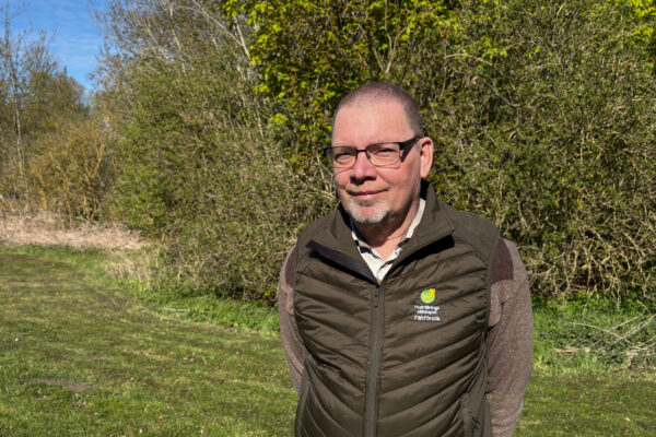 Man med glasögon står och tittar mot kameran i soligt vårväder vid en grönskande buskridå. Han bär grön väst med texten ”Hushållningssällskapet” och ”Fältförsök” samt en skjorta.
