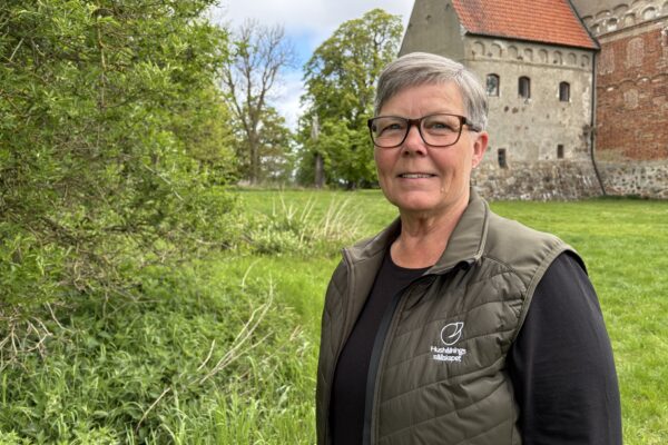 Kvinna med kort grått hår och glasögon står och tittar mot kameran i en grön parkmiljö. Hon bär väst med texten ”Hushållningssällskapet”, och bakom syns ett äldre stenhus med rött tegeltak.