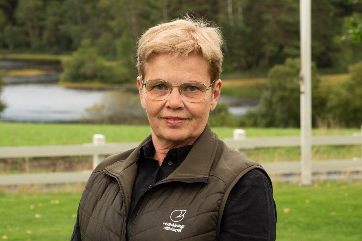 Porträtt av en kortklippt kvinna med glasögon som står utomhus och ser in i kameran, klädd i mörk skjorta och väst med texten ”Hushållningssällskapet”, med sjö, gräs och skog i bakgrunden.
