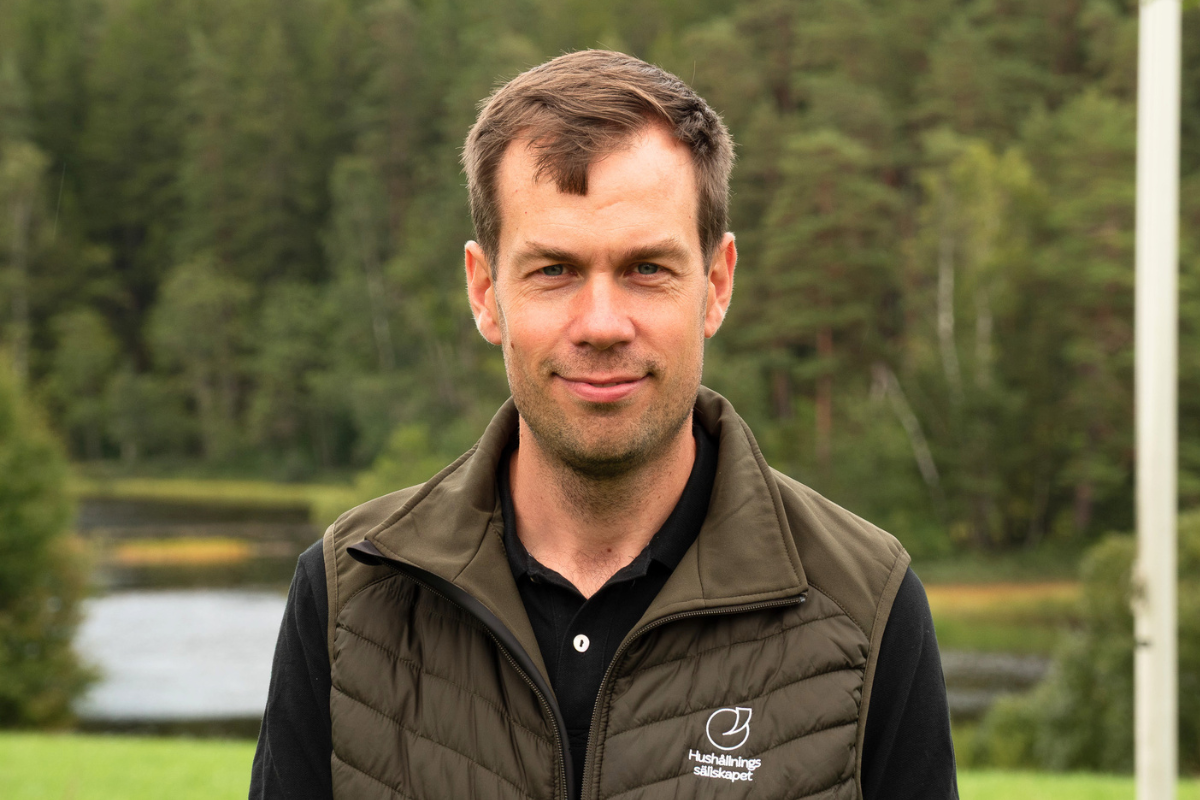 Man med kort brunt hår står utomhus och ser in i kameran med ett lätt leende, iklädd mörk piké och quiltad väst med loggan och texten ”Hushållningssällskapet”, med skog och vatten i suddig bakgrund.