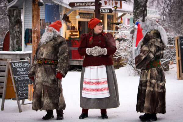 Tre personer i vinterkläder står och pratar på en snötäckt julmarknad; två i långa pälsar och röda tomteluvor, en i folkdräkt med stickade vantar. Skyltar visar “BANVAKTSTUGAN”, “STADEN”, “KRETSLOPPSHUSET” och “GLÖGG MUGG 40:-”.