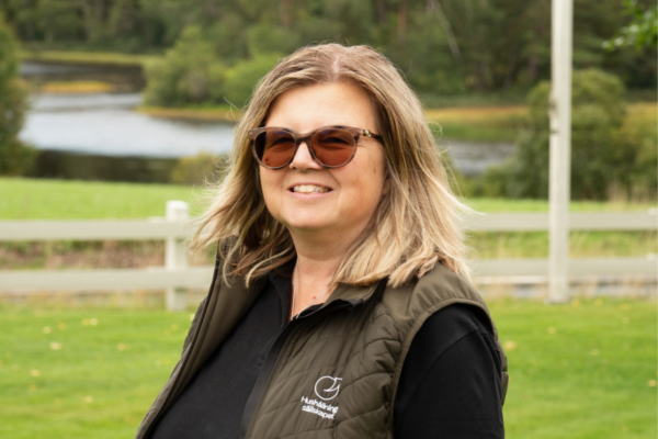Leende kvinna med solglasögon och axellångt hår står utomhus på en grön gräsmatta. Hon bär mörk tröja och väst med loggan och texten ”Hushållningssällskapet”, med sjö och träd i bakgrunden.