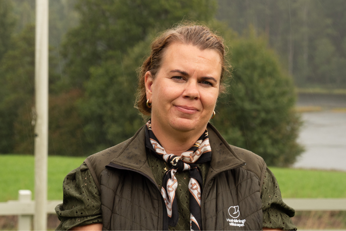 Kvinna med uppsatt hår och diskret leende står utomhus i lätt regn, klädd i mörk väst och mönstrad scarf. På västen syns logotypen och texten ”Hushållningssällskapet”; grönskande bakgrund med vatten.