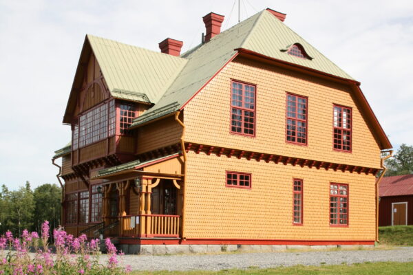 Stor gul träbyggnad med grönt plåttak och röda fönsterkarmar står i solljus, med veranda och inglasad del på gaveln. I förgrunden växer höga rosa blommor på en äng intill grusplan.