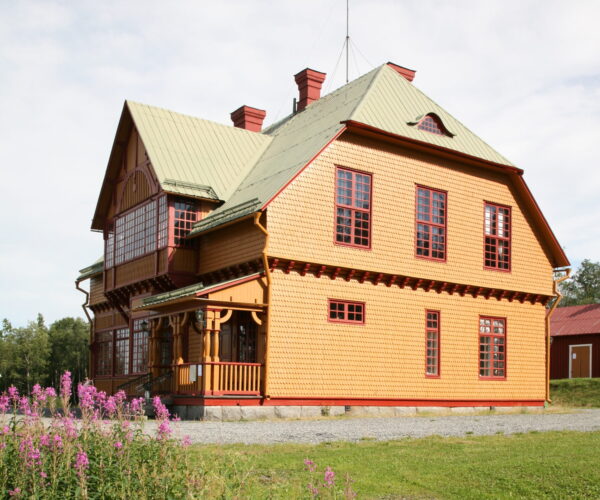 Stor gul träbyggnad med grönt plåttak och röda fönsterkarmar står i solljus, med veranda och inglasad del på gaveln. I förgrunden växer höga rosa blommor på en äng intill grusplan.