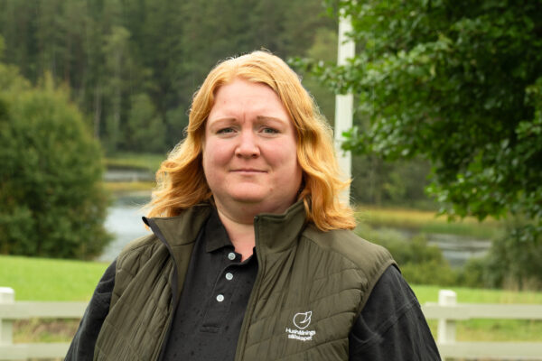 Kvinna med rött hår står och tittar in i kameran utomhus i lätt regn, med skog och vattendrag i bakgrunden. Hon bär grön väst med texten “Hushållningssällskapet” över en svart piké.