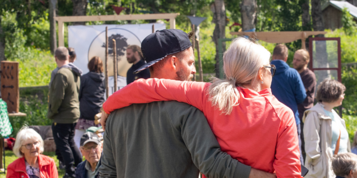 Ett par står med armarna om varandra och tittar ut över en folksamling. Runt dem syns besökare som pratar och rör sig mellan skyltar och aktiviteter i en grön parkmiljö i dagsljus.