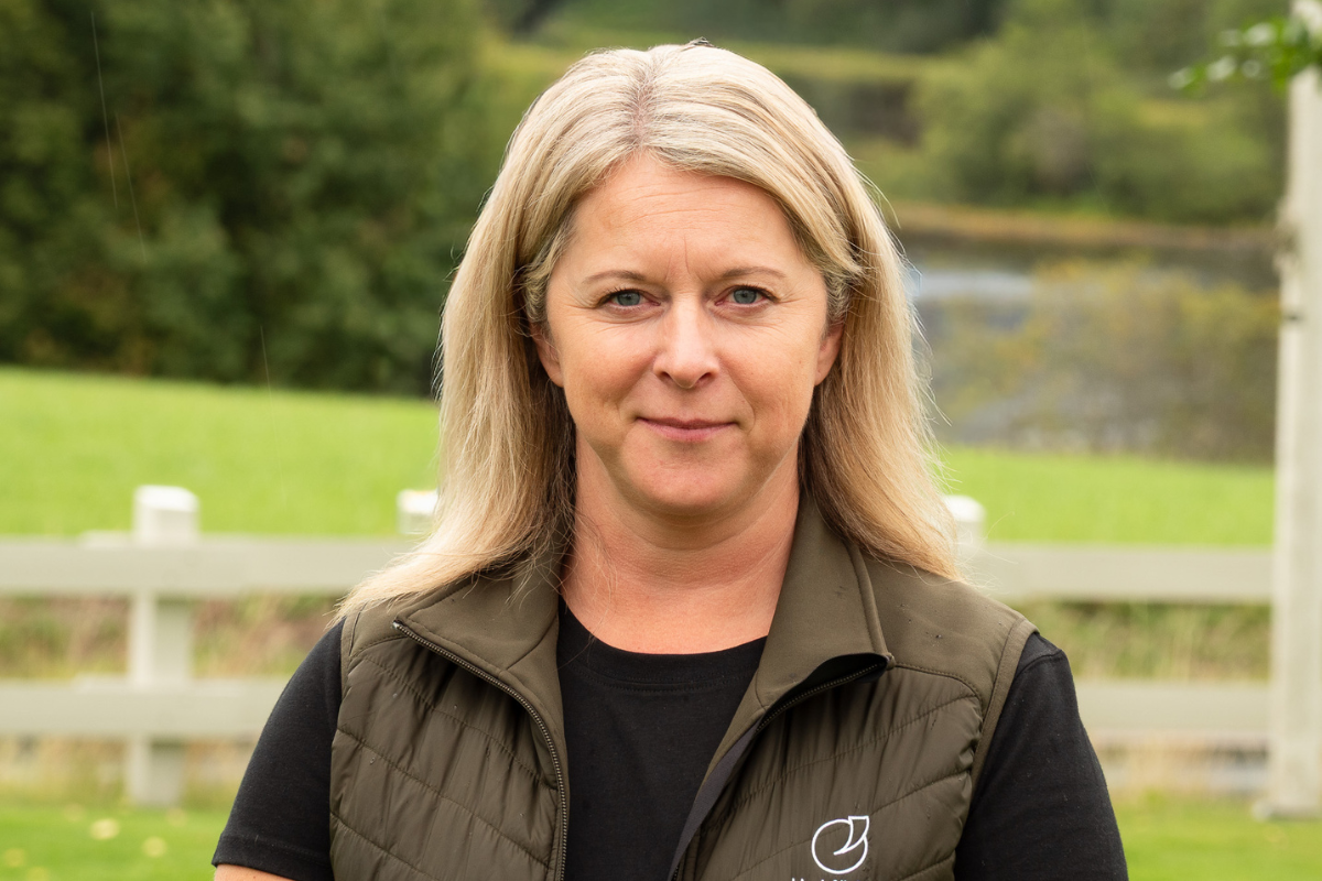 Blond kvinna står utomhus och ser in i kameran med ett neutralt ansiktsuttryck. Hon bär svart tröja och grön vadderad väst med liten vit logotyp, med grön äng, vitt staket och träd i bakgrunden.