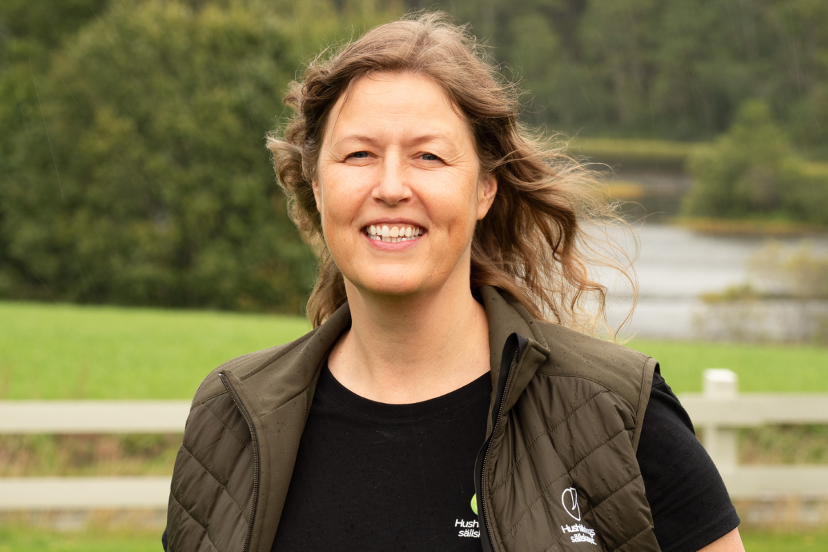 Leende kvinna står utomhus och tittar mot kameran, med vind som blåser i håret. Hon bär svart tröja och mörkgrön quiltad väst med texten “Hushållsällskapet”, framför grönskande landskap och vatten.