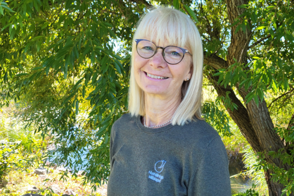 Leende kvinna med blont hår och glasögon står utomhus och tittar mot kameran. Hon bär en mörk tröja med loggan och texten ”Hushållnings sällskapet”, och står framför ett lummigt träd vid vatten.