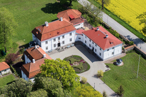 Flygfoto visar en vit herrgårdsliknande byggnad med röda tegeltak som omger en innergård med rondell och planteringar. Huset ligger vid grönskande park, åkrar och en landsväg i bakgrunden.