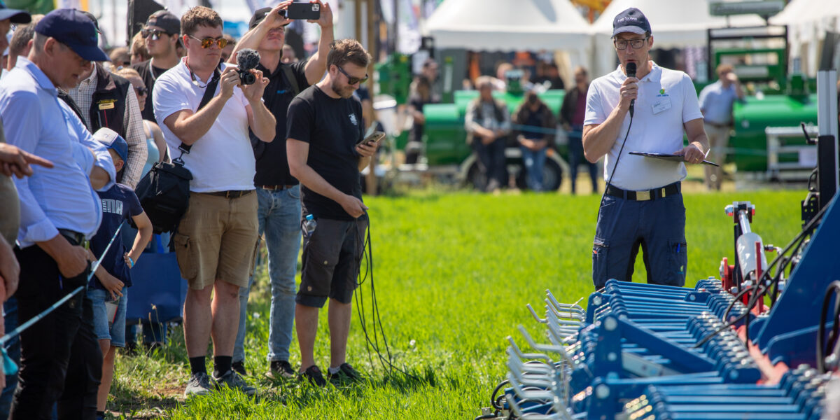 Man i vit pikétröja och keps håller mikrofon och läser från en pärm medan han presenterar en blå jordbruksmaskin på en mässa utomhus. Besökare står i gräset, filmar och fotograferar; tält och banderoller syns i bakgrunden.