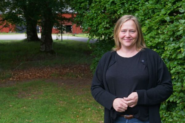 Smiling woman with shoulder-length blond hair stands with hands clasped beside dense green bushes, wearing a black cardigan and top. Background shows a grassy area, trees, and a red building across a quiet road.