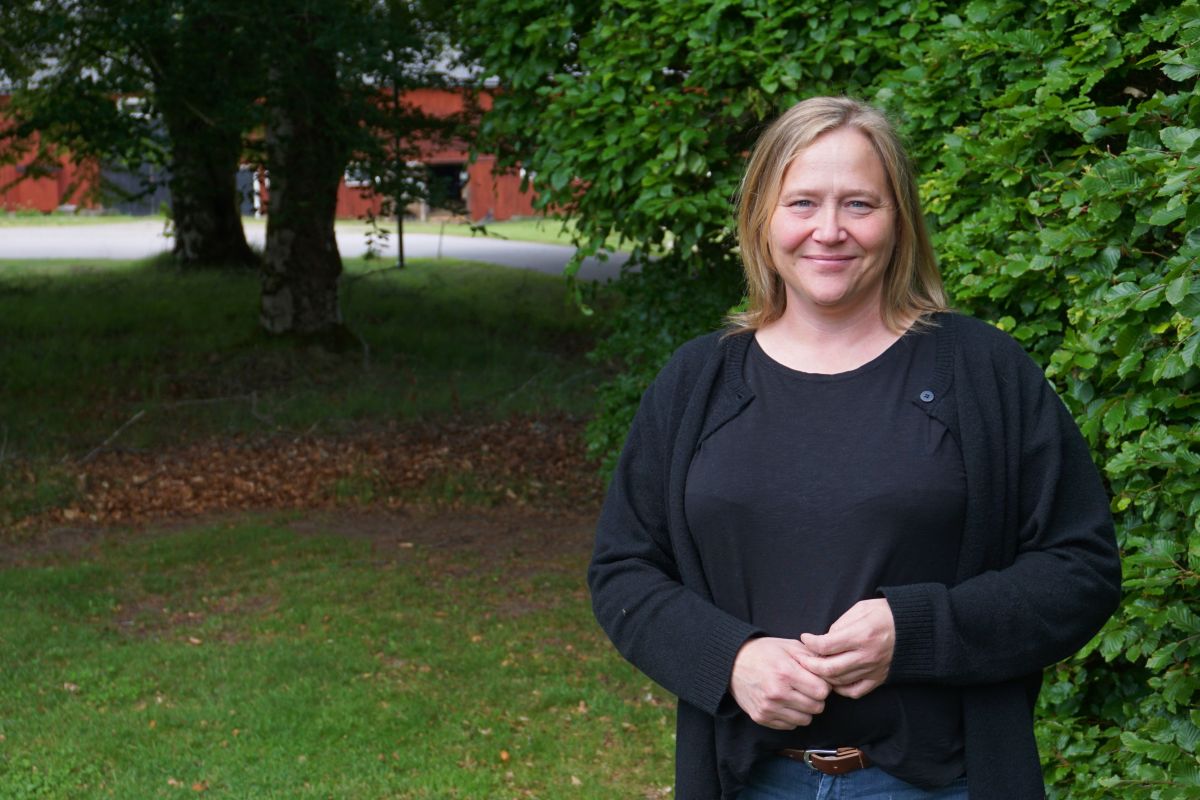 Leende kvinna med axellångt blont hår står med händerna samlade framför sig, klädd i svart tröja och kofta, vid en tät grön häck. I bakgrunden syns gräsmatta, träd och en röd byggnad.