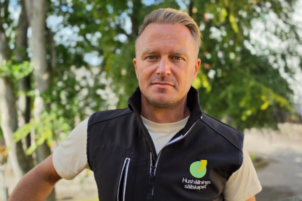 Man med kort, ljusbrunt hår står utomhus och ser in i kameran, med ena armen i sidan. Han bär svart väst med logotyp och texten ”Hushållnings sällskapet”, mot suddig bakgrund av gröna träd.
