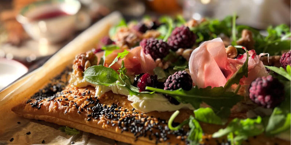Långt frasigt smördegsflarn toppas med färska björnbär, grön sallad och örter, nötter och tunna skivor prosciutto. Rätten ligger på en plåt med bakplåtspapper, med julig serveringskänsla i varm belysning.