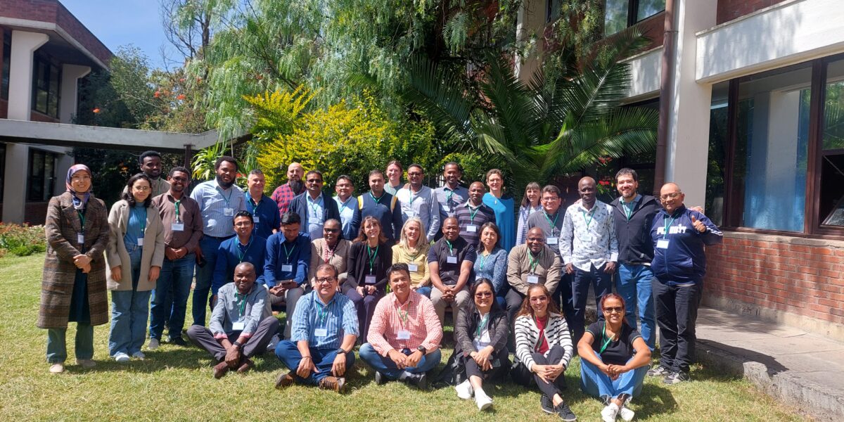 Stor grupp människor poserar och ler för ett gruppfoto på en gräsmatta, några sittande längst fram, med namnskyltar runt halsen. I bakgrunden syns frodiga träd och en tegelbyggnad med stora fönster i solsken.