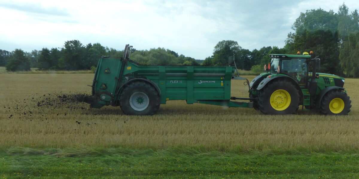Grön traktor drar en gödselspridare och sprider mörk gödsel över en skördad åker. Spridaren är märkt “FLEX 16” och “Samson”. Trädlinje och grå himmel syns i bakgrunden.