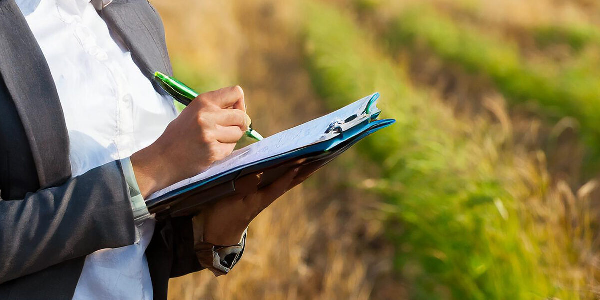 Person i kavaj skriver med grön penna på ett block i en clipboard, som hålls i handen. Fotot är taget utomhus med suddig bakgrund av gräs och fält, som vid en inspektion eller kontroll.