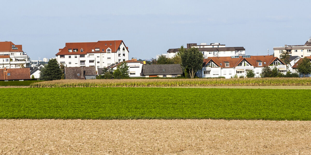 Bostadshus och flerfamiljshus ligger på rad i utkanten av en stad, bakom stora åkrar. I förgrunden syns ett plöjt fält och ett brett grönt odlingsfält, under klar blå himmel.