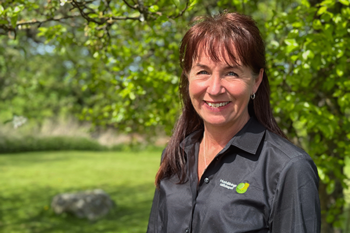 Jenny Fredman står utomhus framför ett grönt, lövklätt träd i solsken. Hon har mörkt, axellångt hår med lugg och bär en svart skjorta med Hushållningssällskapets logotyp. Hon ler varmt mot kameran, och i bakgrunden syns en grön gräsmatta och en sten.