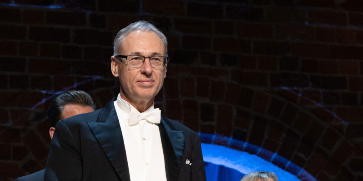 Man i frack och vit fluga står på en scen och ser framåt, som vid en högtidlig ceremoni eller konsert. Bakom honom skymtar andra personer och en mörk sal med blå belyst båge.