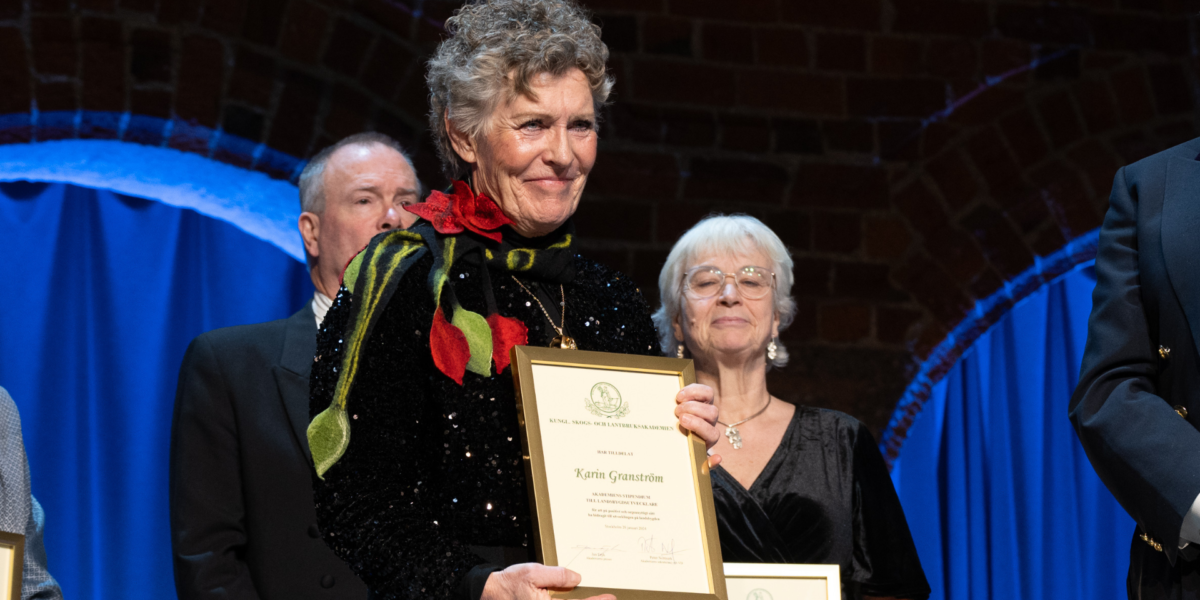 Kvinna på scen håller upp ett inramat diplom och ler, omgiven av andra deltagare vid en ceremoni. Text på diplomet: “KUNGL. SKOGS- OCH LANTBRUKSAKADEMIEN … Karin Granström”. Blå ridå och mörk bakgrund.