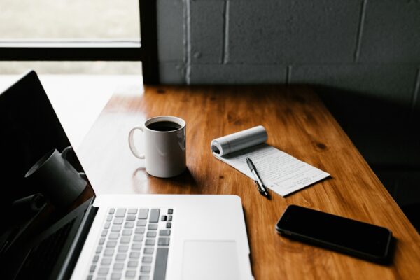 Bärbar dator står öppen på ett träbord bredvid en kopp kaffe, en mobiltelefon och ett block med handskrivna anteckningar och penna, i ett ljust arbetsutrymme nära ett fönster.