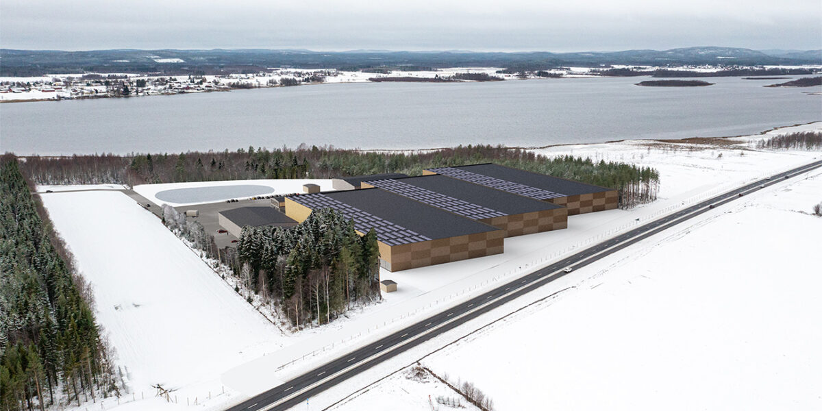 Drönarvy över snötäckt landskap där en stor, rektangulär industribyggnad med mörkt tak och solpaneler är inplacerad som illustration intill en skogsdunge och en väg; i bakgrunden syns en bred sjö och by.