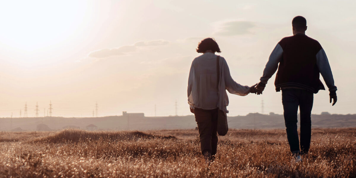 Två personer går hand i hand över ett torrt fält i motljus, sedda bakifrån. Solen står lågt på himlen och kastar varmt ljus över landskapet, med siluetter av elmaster och byggnader i fjärran.