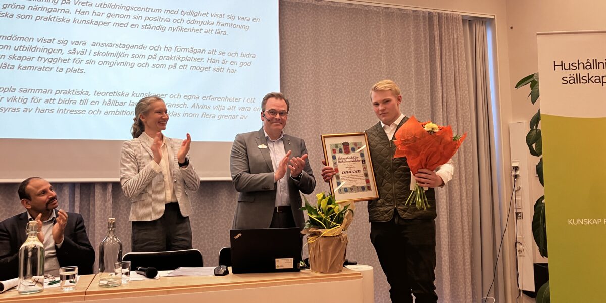 Ung man tar emot diplom och blomsterbukett medan två personer bredvid applåderar vid en prisutdelning. På projektorduken står ”Stipendiat 2023 - motivering”, och en rollup visar texten ”Hushållningssällskapet”.