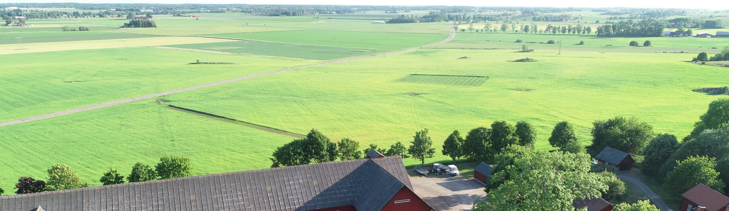 Flygfoto över vidsträckta gröna åkrar där en smal landsväg slingrar sig genom landskapet; i förgrunden syns röda ladugårdsbyggnader, träd och en gårdsplan med parkerade bilar, med skogsridå och gårdar i fjärran.